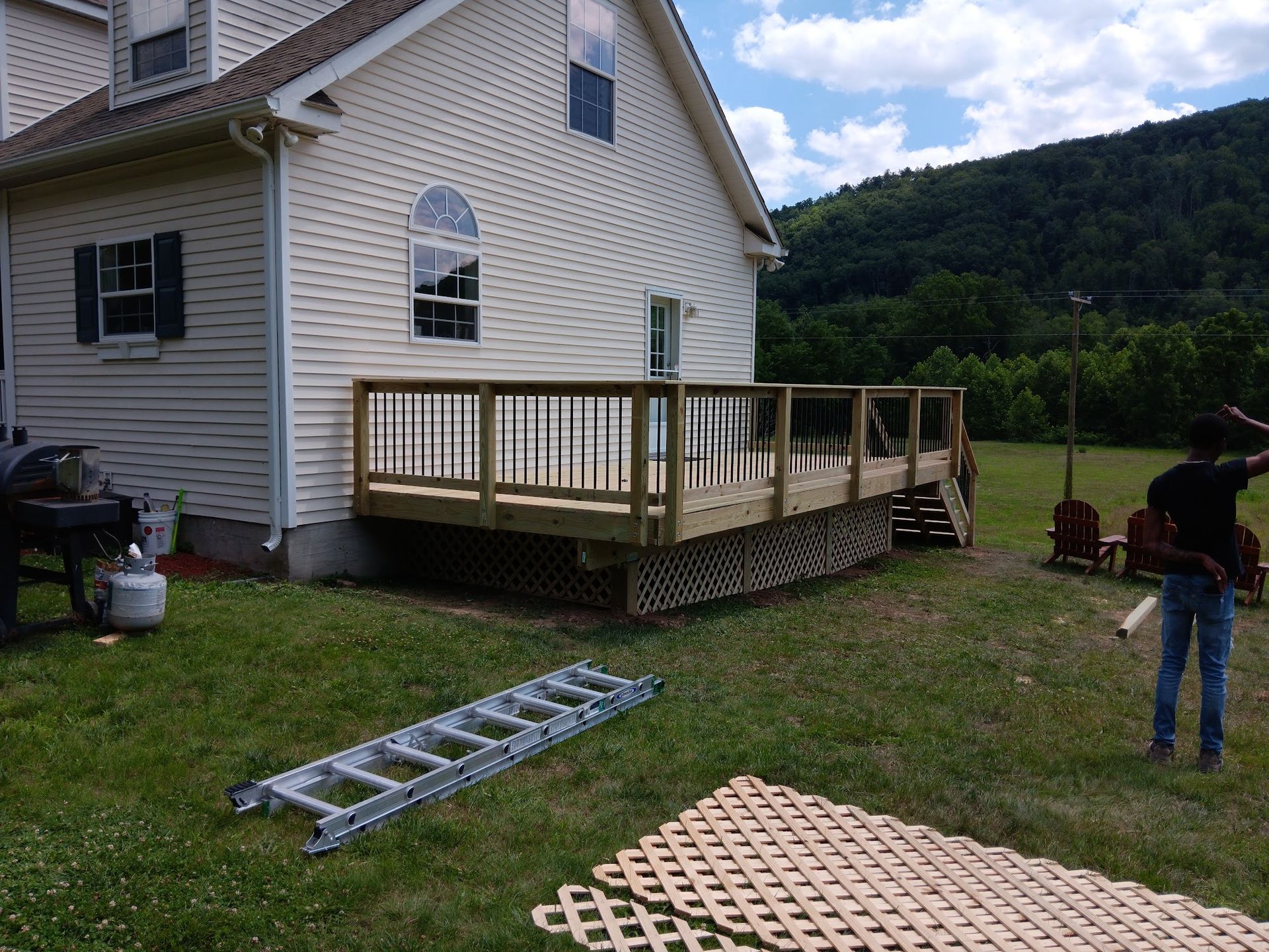 Backyard deck attached to a house; person holding lumber; ladder and lattice in foreground; trees in background.