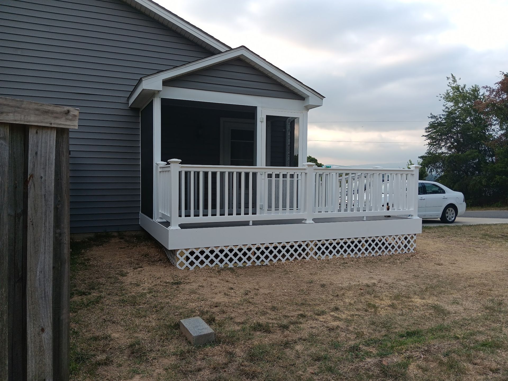 Screened-in porch attached to a gray house, with white railings and lattice, surrounded by dry grass.