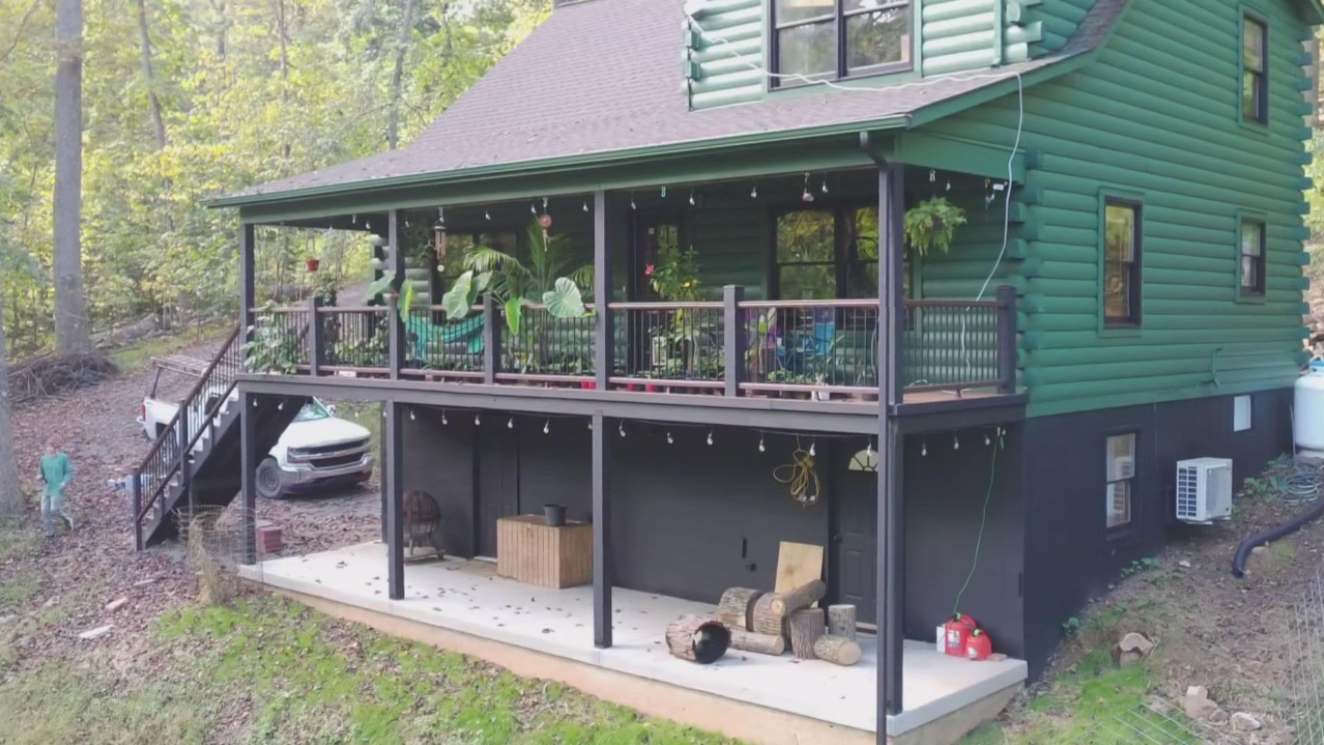 Green cabin with a deck, string lights, and a lower-level concrete patio with firewood.