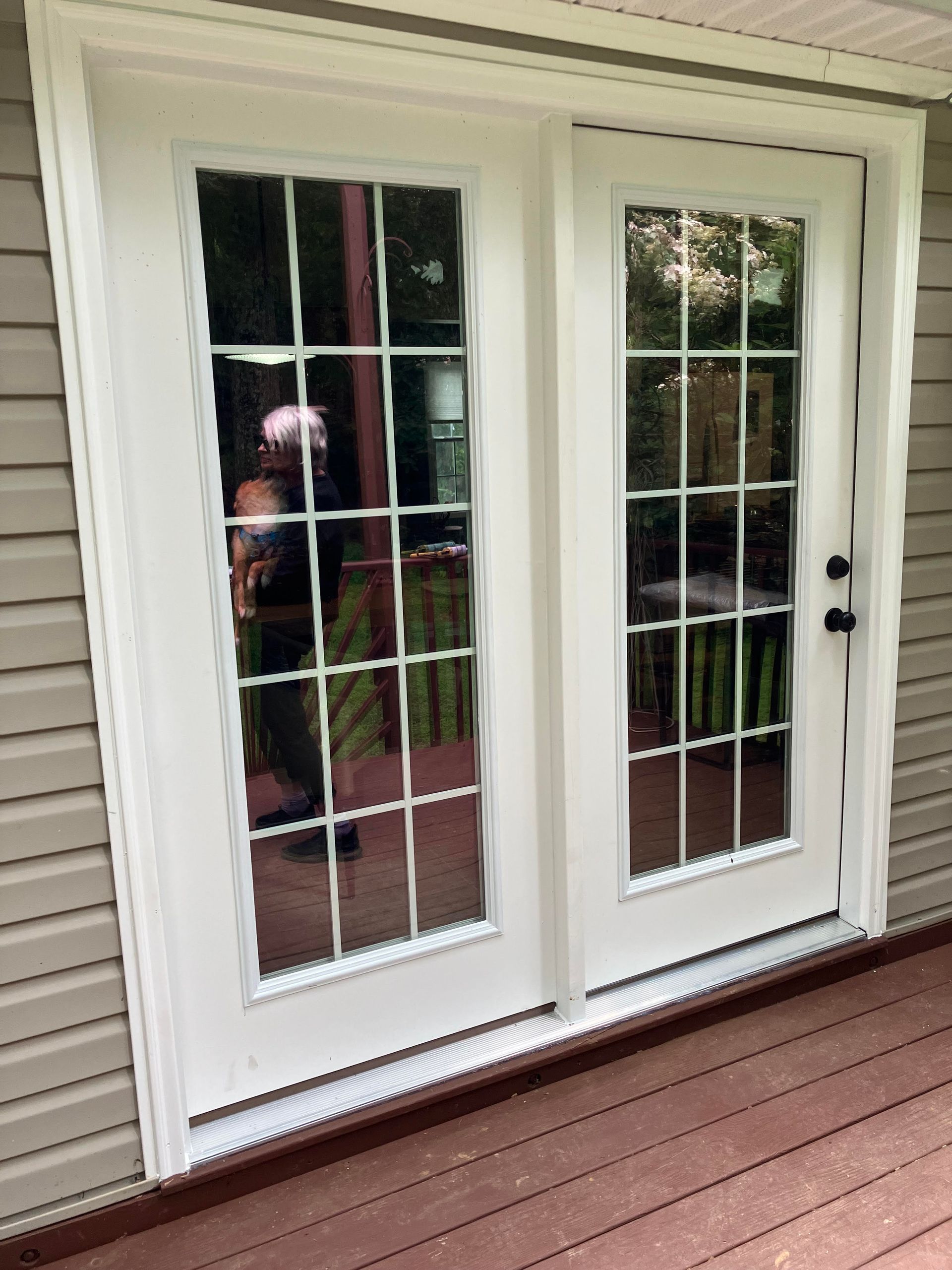 White French doors with multiple window panes, set in a wood frame on a deck, with a person reflected in the glass.