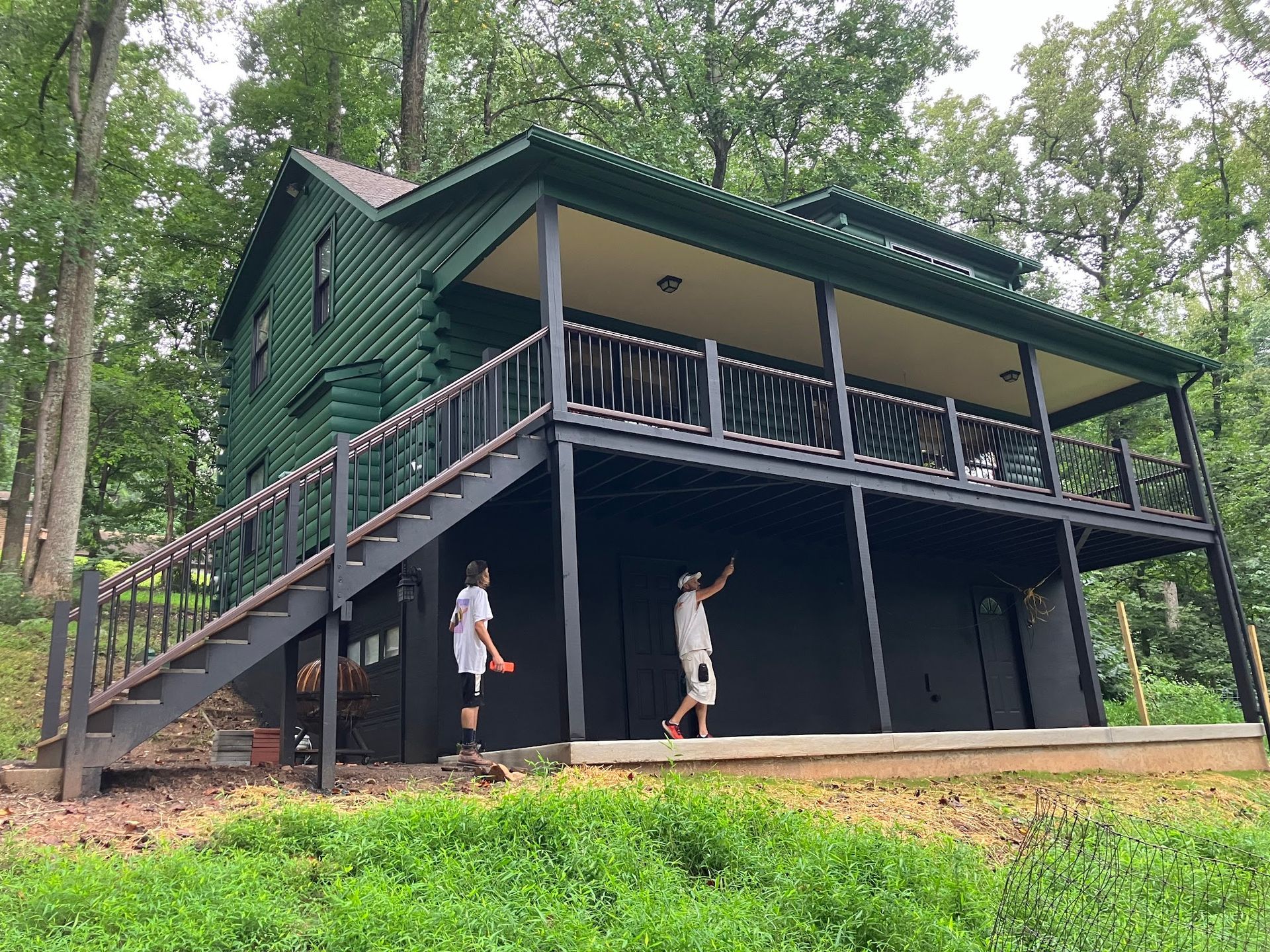 Green house with black trim, a deck, and stairs. Two people stand below on concrete.