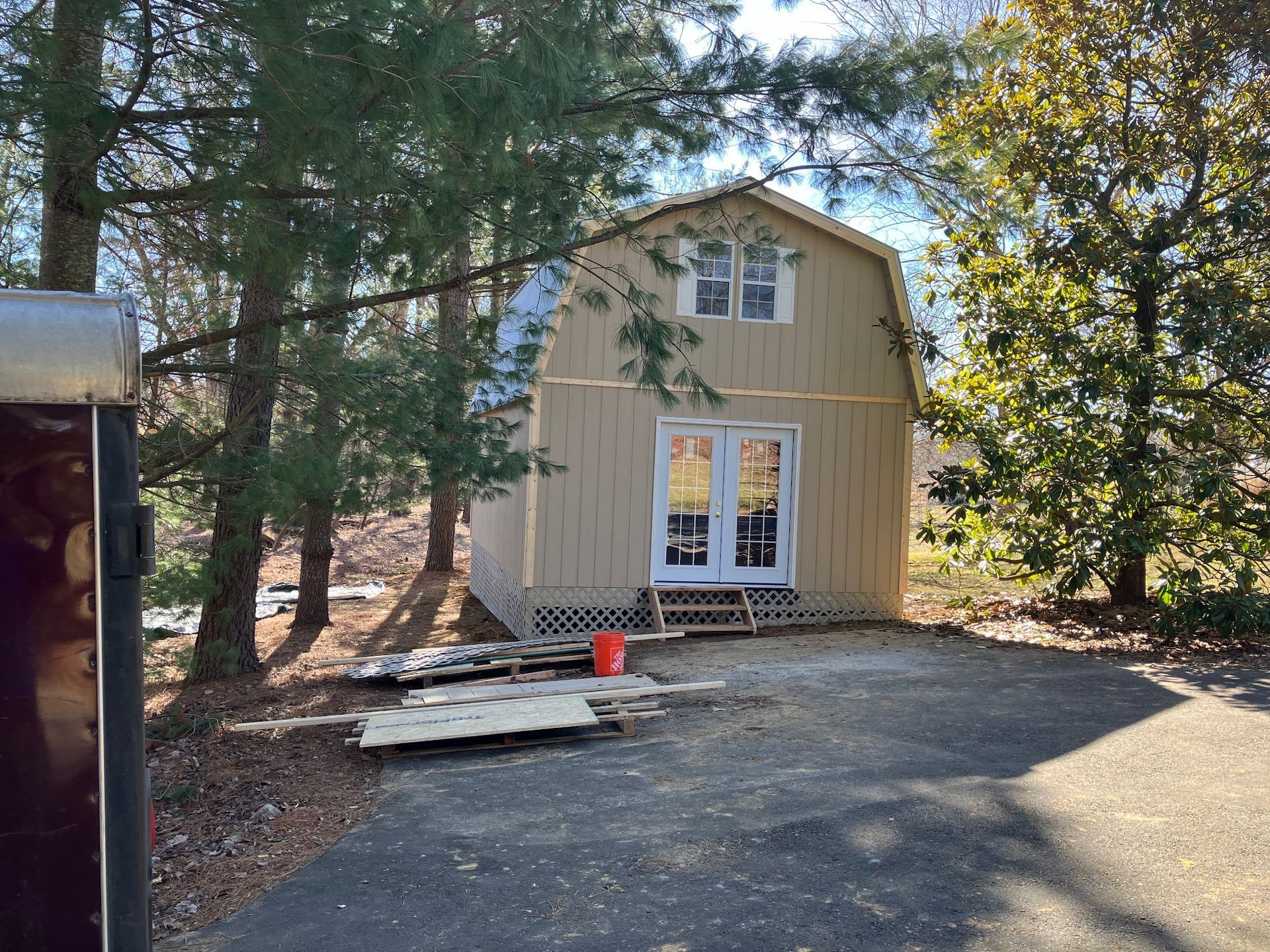 Tan barn-shaped shed with double doors and small upper window, surrounded by trees and a driveway.