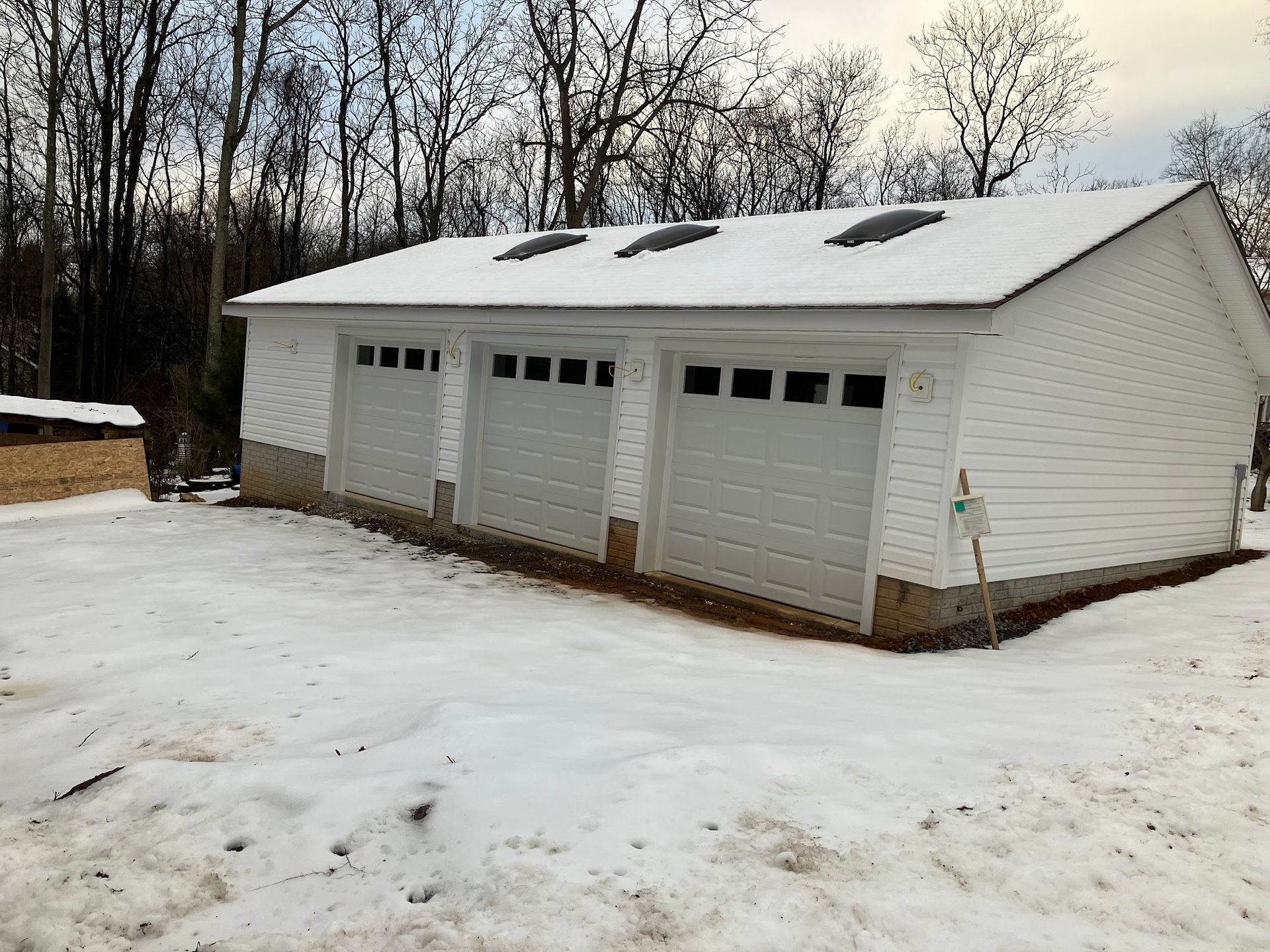 White three-car garage in snow, with three garage doors and roof skylights. Trees in the background.