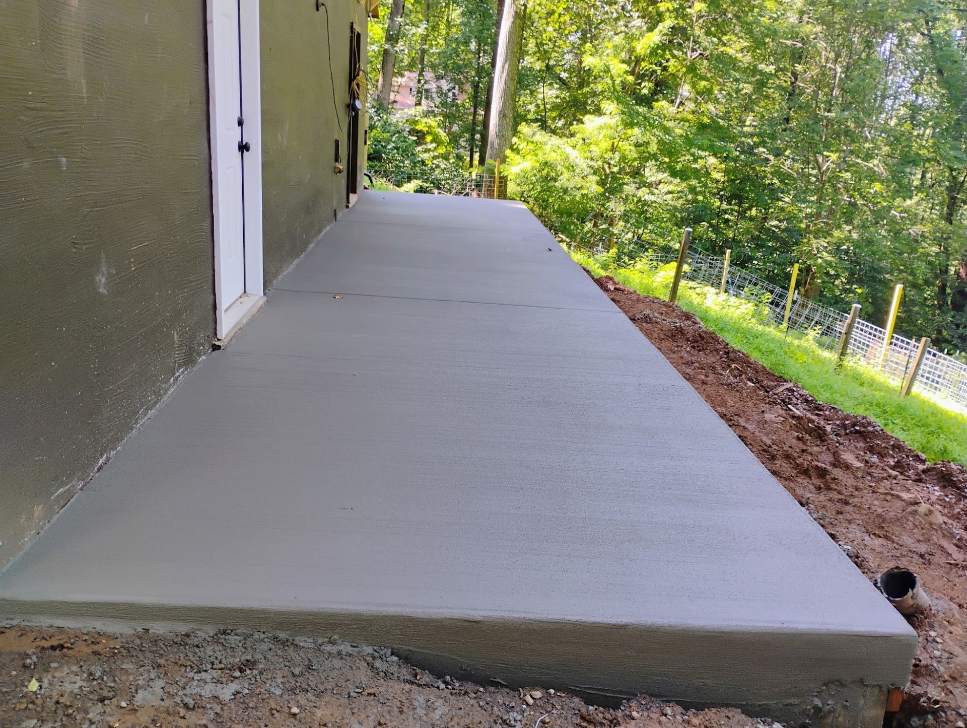 Newly poured concrete patio next to a house with a door, overlooking a grassy hill and trees.
