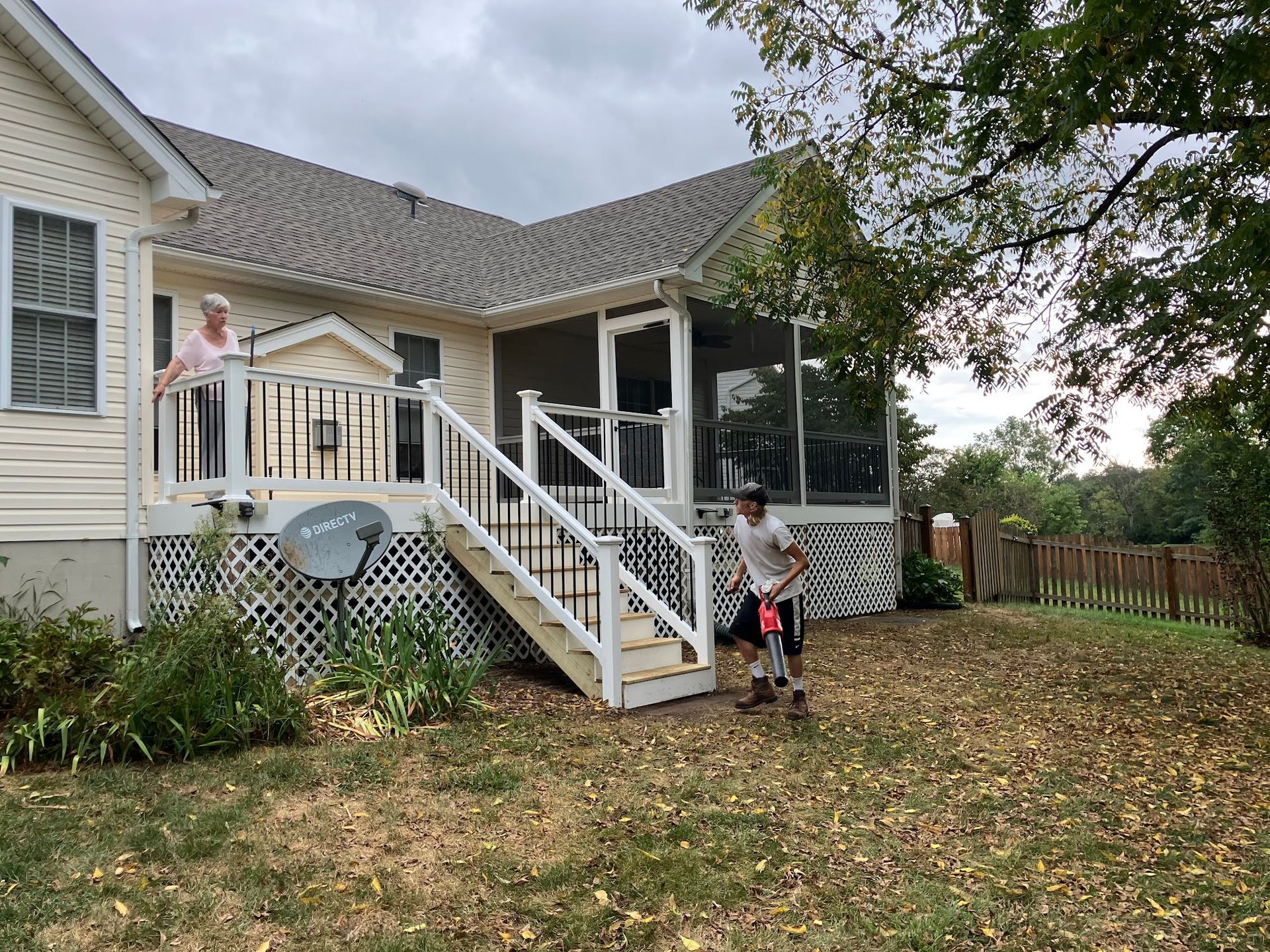 A person rakes fallen leaves in a yard with a deck and screened porch. Another person stands on the deck.