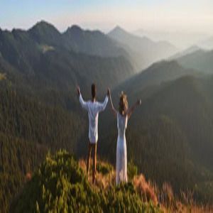 Two people standing on a mountain overlook with arms outstretched at sunrise over forested hills