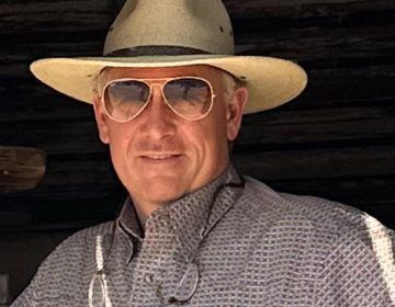 A man wearing a cowboy hat and sunglasses is smiling.