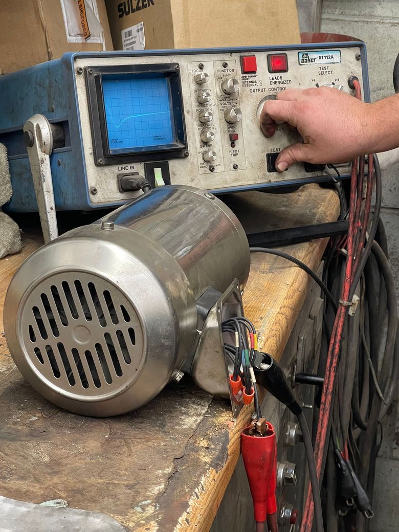 A hand adjusts the dial on an oscilloscope connected to a cylindrical metal electric motor via jumper cables. A hand adjusts the dial on an oscilloscope connected to a cylindrical metal electric motor via jumper cables.