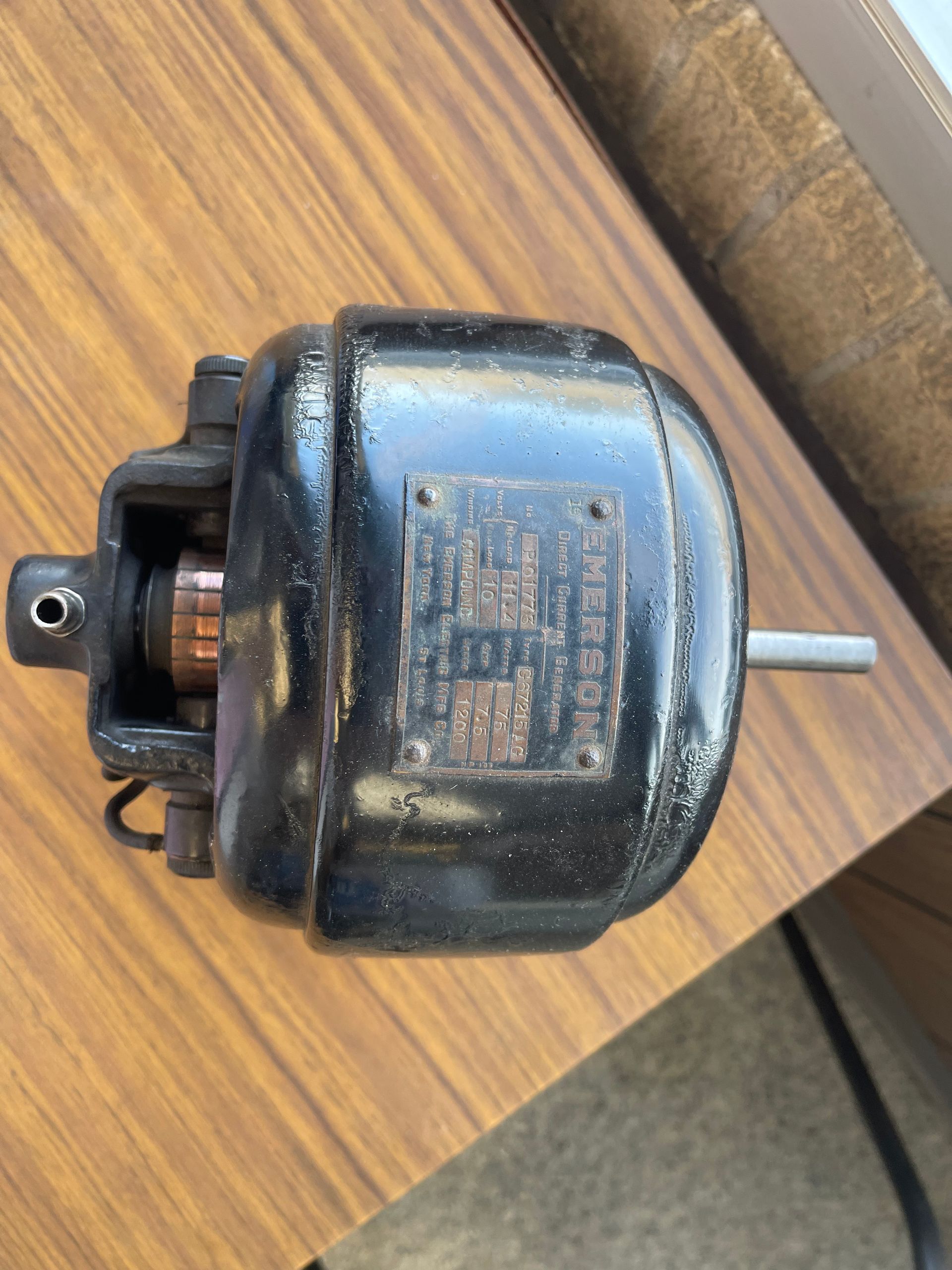 A small, black electric motor sits on a wooden table, showing a copper commutator and a metal information plate.