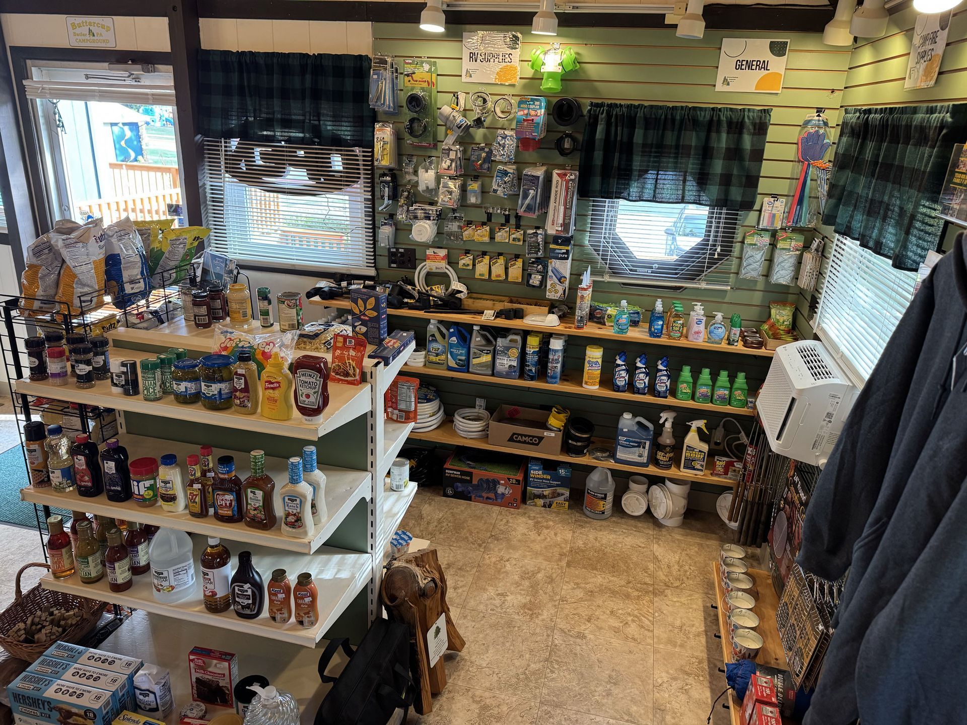 A retail shop interior with shelves stocked with products, green walls, and two windows with plaid curtains.