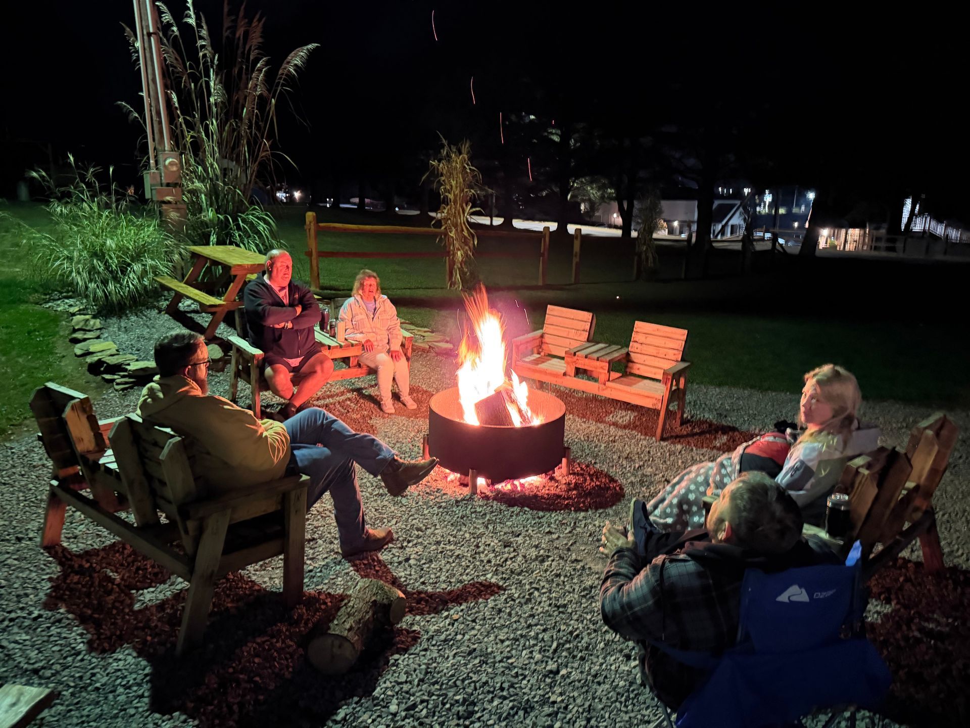 People seated around a fire pit at night, chatting. Wooden benches on gravel.