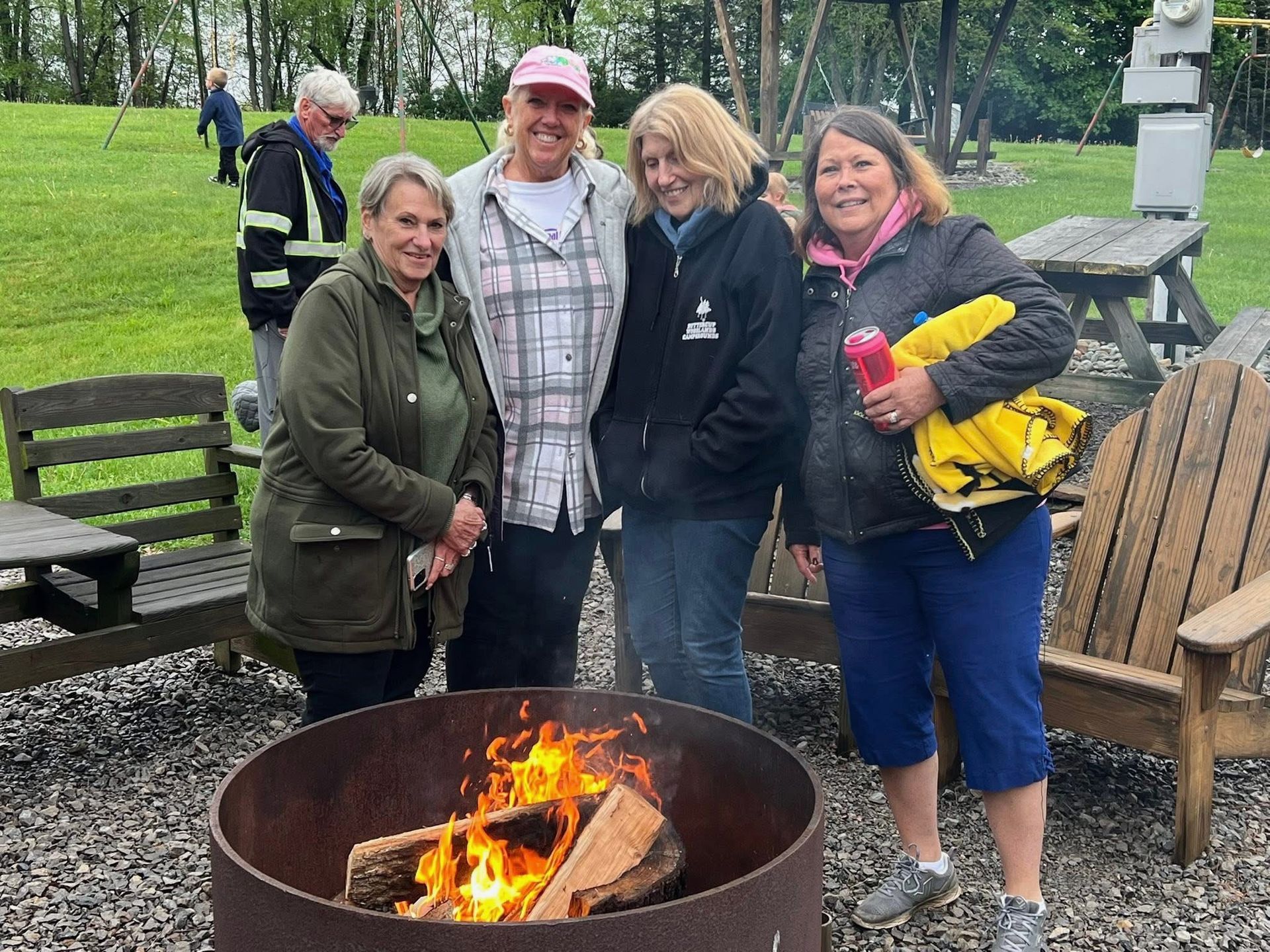 Four women standing around a campfire. Outdoors, near wooden chairs and a grassy area.
