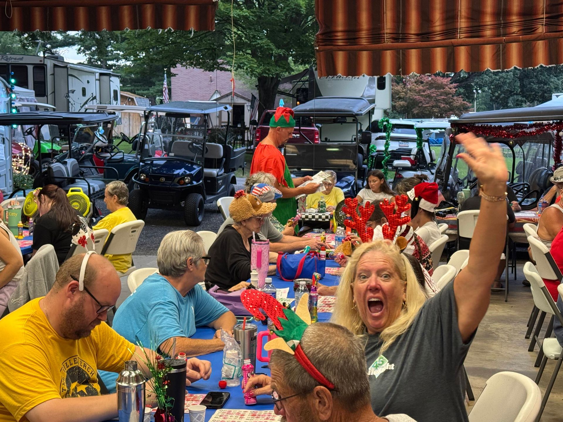 People at outdoor Christmas-themed party; waving, wearing festive headbands. Tables set; golf carts in the background.