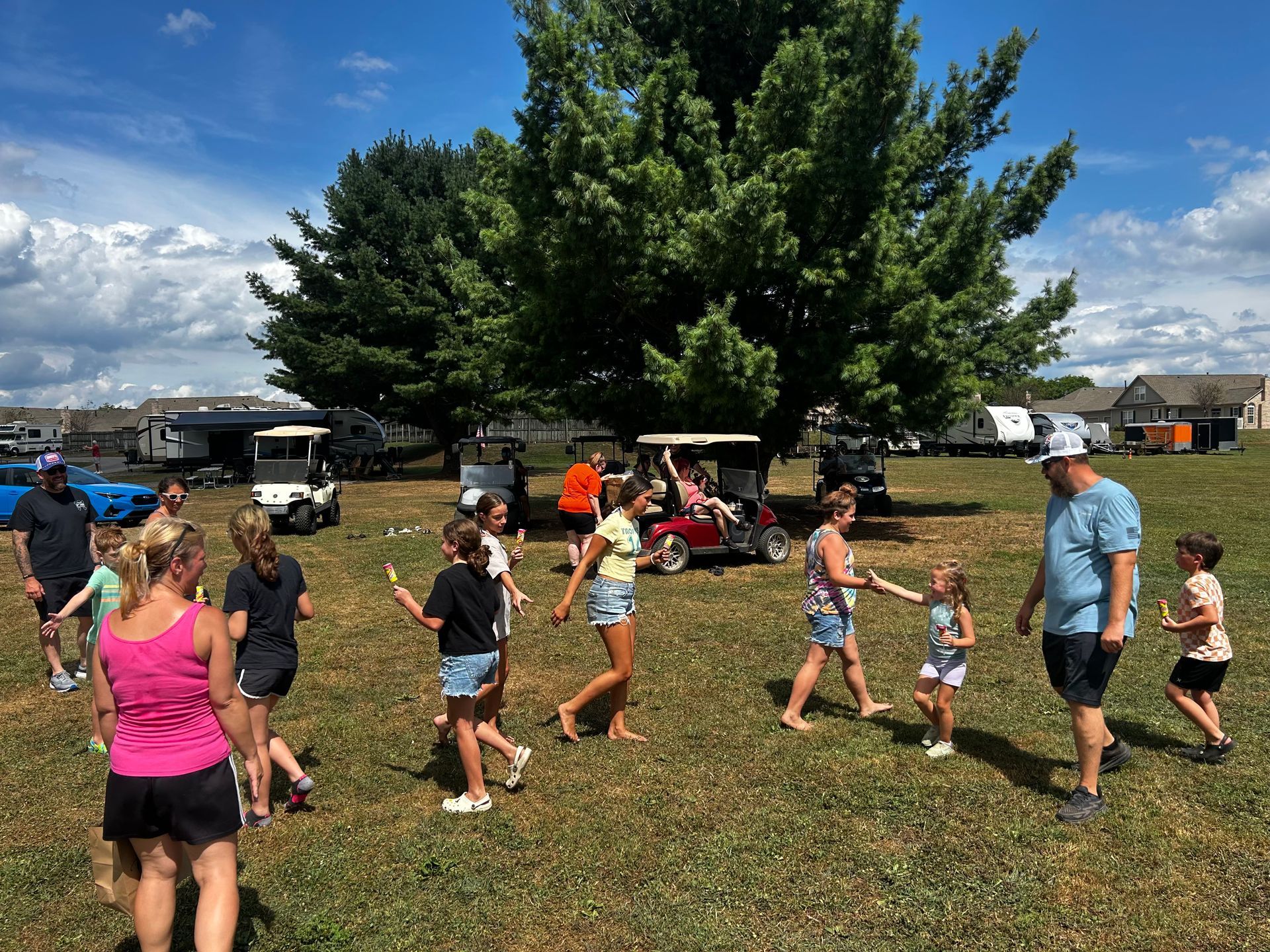 People playing in a grassy field with golf carts and large trees on a sunny day.