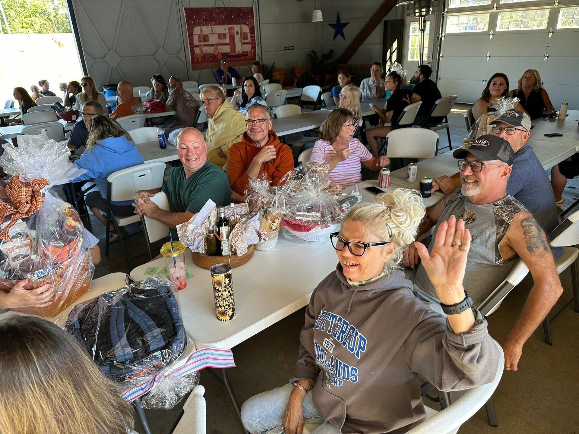 People at tables in an open-air building. Some wave, gift baskets on the tables.