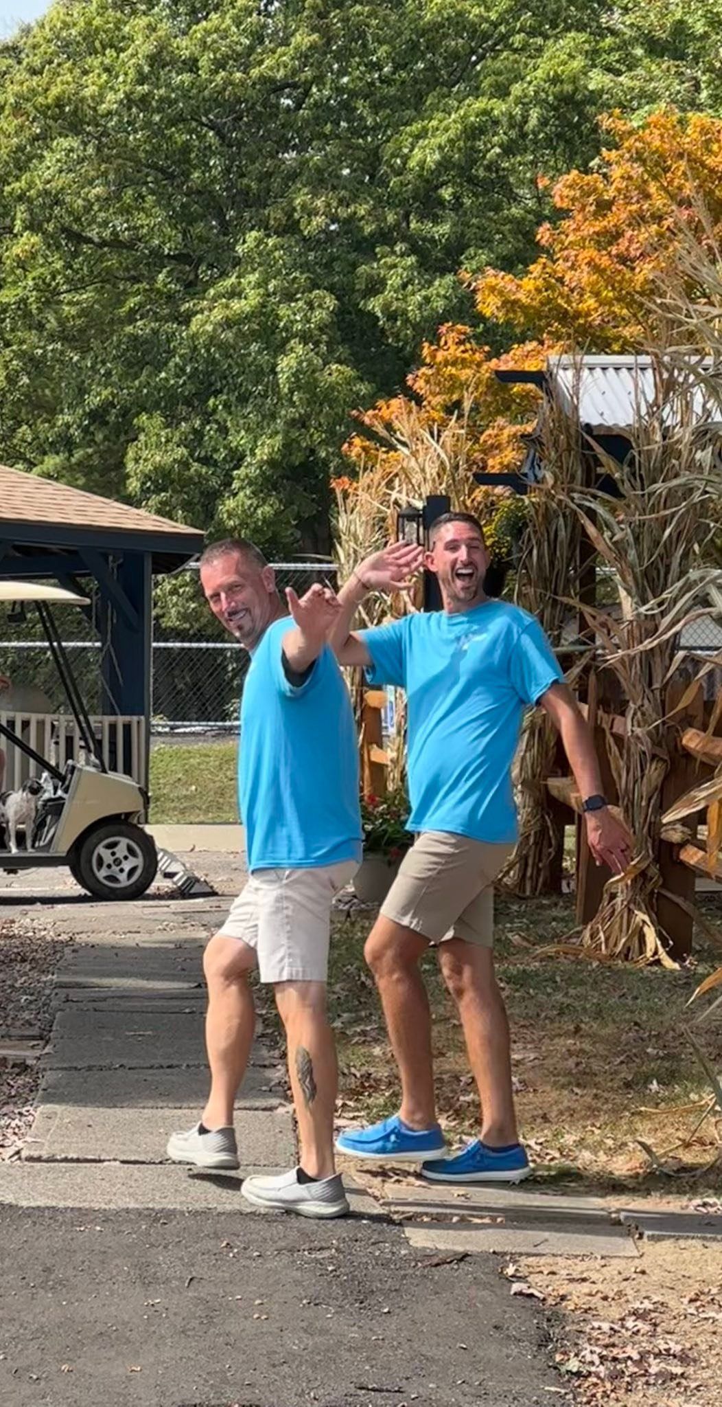 Two men in blue shirts and tan shorts wave excitedly outdoors.