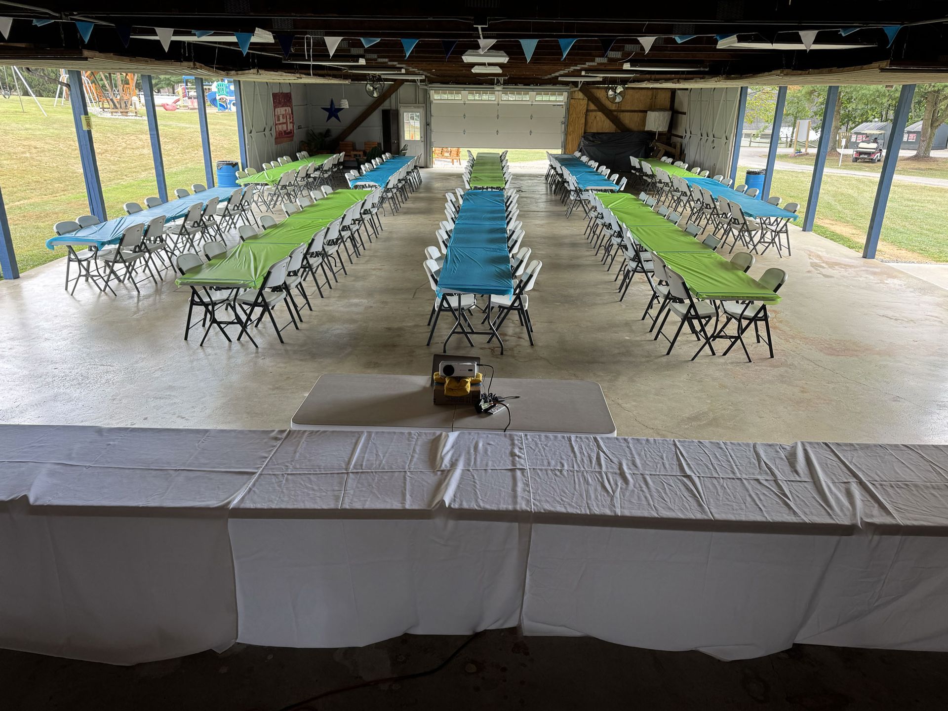 Long tables covered in blue and green cloths set up under a covered outdoor space; a speaker sits on a stage.