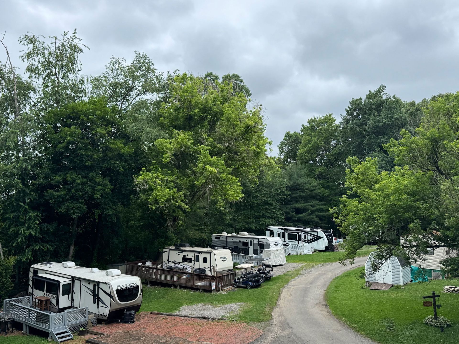 RVs parked in a wooded campground on a cloudy day. A gravel road leads to the sites.