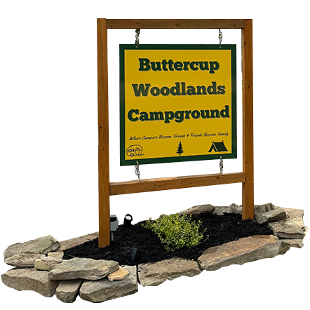 Sign for Buttercup Woodlands Campground, brown frame, green and yellow sign, stones, black mulch, small green plant.