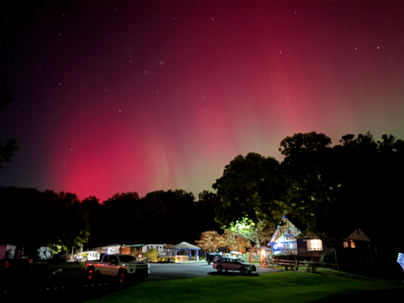 Night sky with pink aurora borealis over a camping area with cars, buildings, and trees.