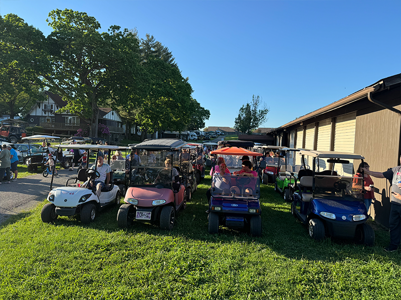 Golf carts parked on grass, people inside. Building and trees in the background, blue sky.