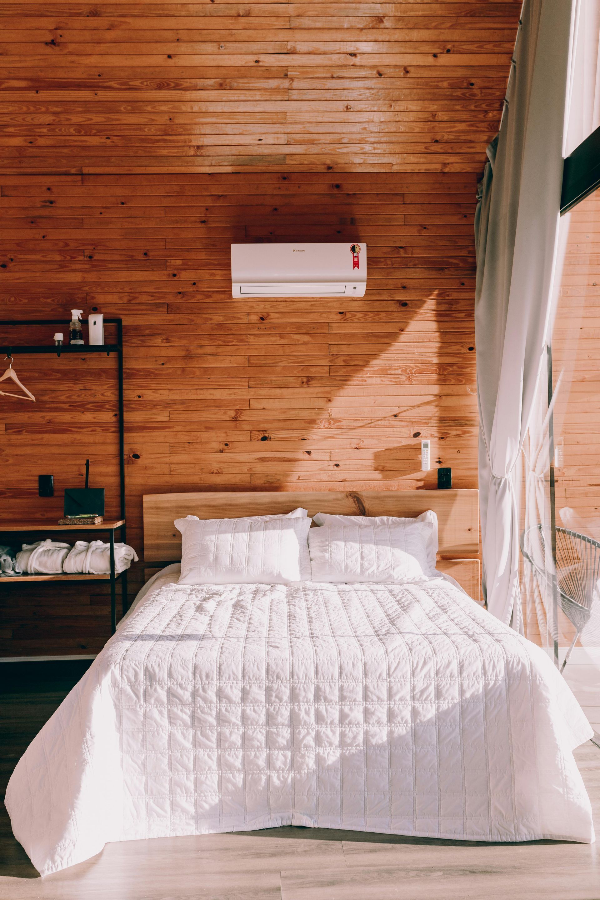 Bedroom with white bedding, wooden walls, air conditioner, and sunlight streaming in.