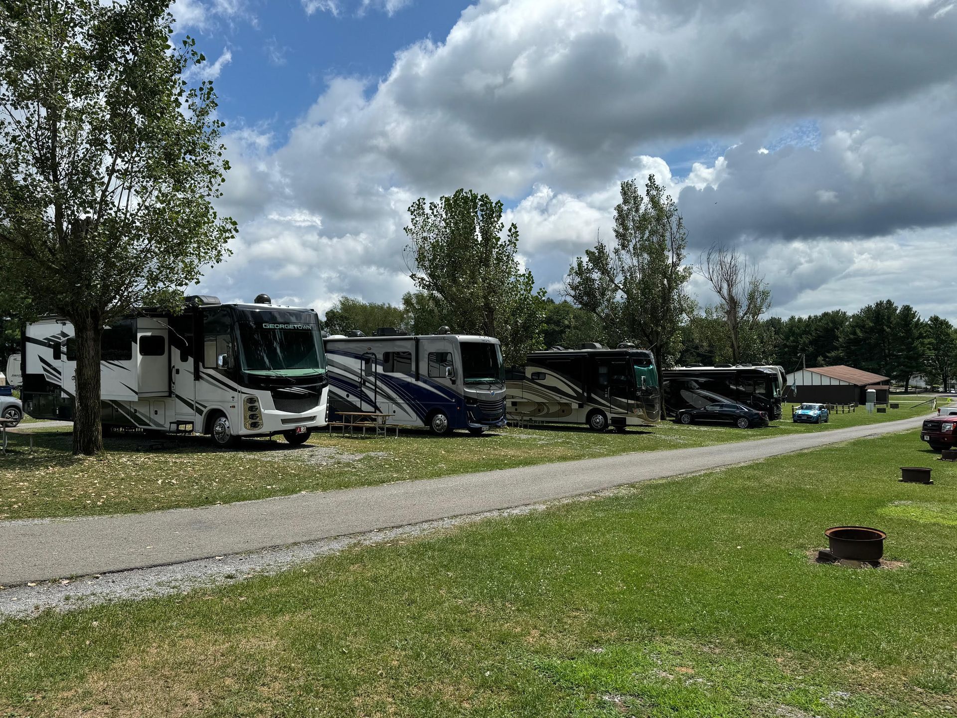 RVs parked in a grassy campground. Gravel path in foreground, trees and cloudy sky in background.