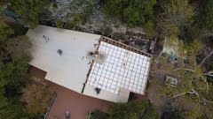 Aerial view of a roof being constructed, partially covered with white panels. Surrounded by trees.