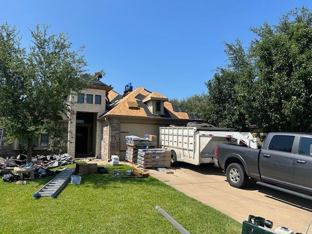 Roofing work in progress on a two-story house; truck, trailer, and materials in the driveway.