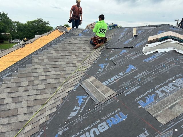 Two roofers working on a roof, installing shingles and underlayment on an overcast day.