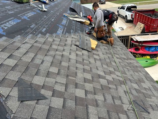 Two workers on a roof, installing asphalt shingles. House exterior, sky visible.