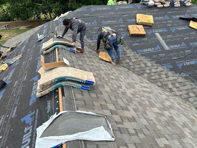 Two roofers installing shingles on a house roof, wearing safety harnesses.