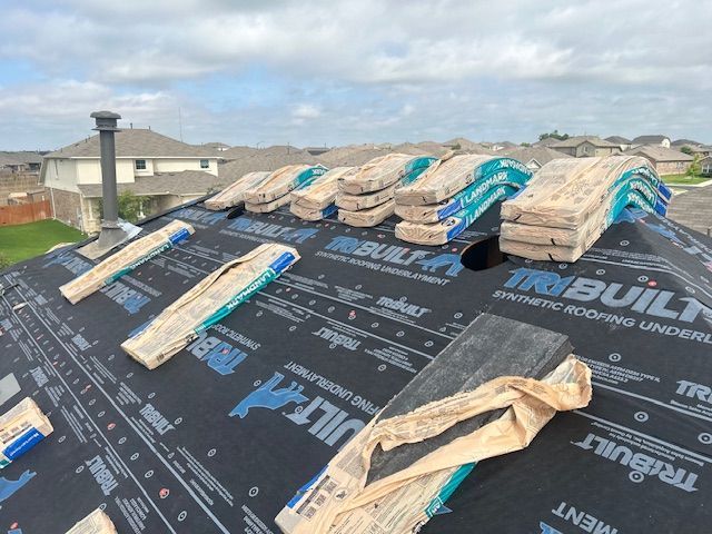 Roof being re-shingled with stacks of shingles on tar paper. Suburban houses and sky are in the background.