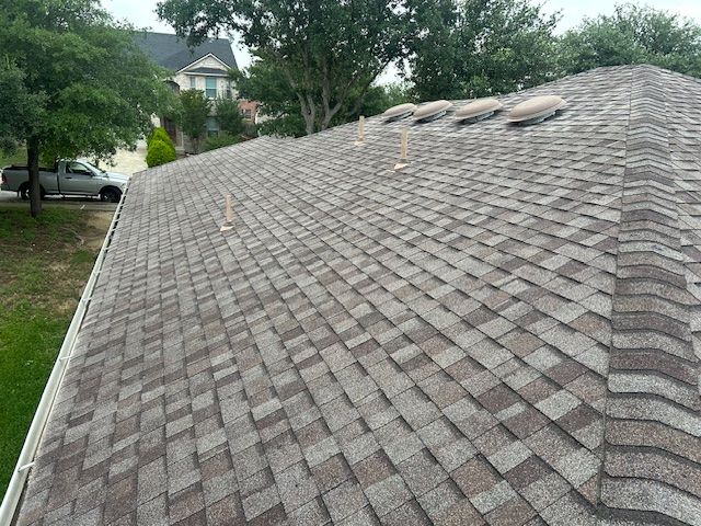 Workers on roof of brick house, replacing shingles. Patio with seating, green lawn, clear sky.