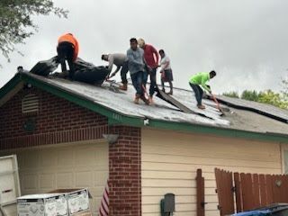 Workers repairing a roof; several people on the roof, using tools. Cloudy sky, brick and siding house.