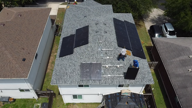 Aerial view of a roof with solar panel installation. Two workers in blue and white stand on the roof, installing panels.