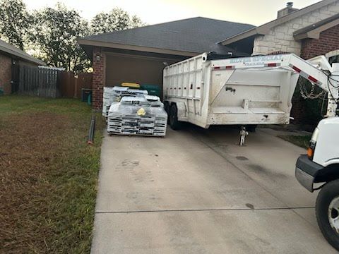A trailer is parked in a driveway next to a house with stacked construction materials. A pickup truck is in view.