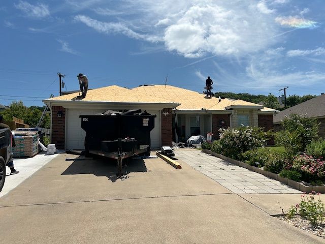 Workers on a roof during a sunny day. A dumpster and trailer are in the driveway.