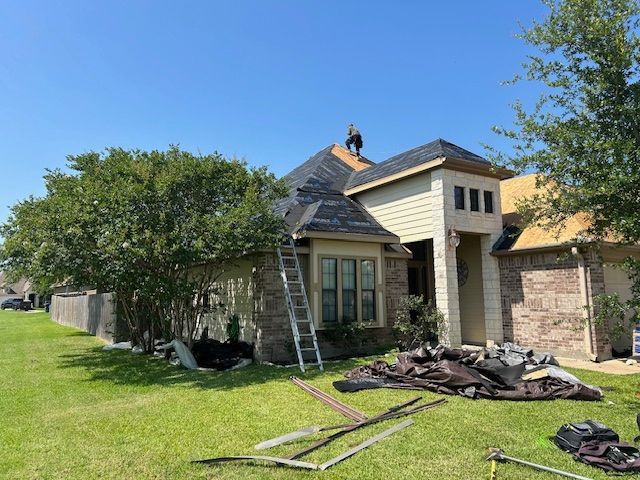 House with roof being repaired; worker on roof, ladder, debris on lawn, sunny day.