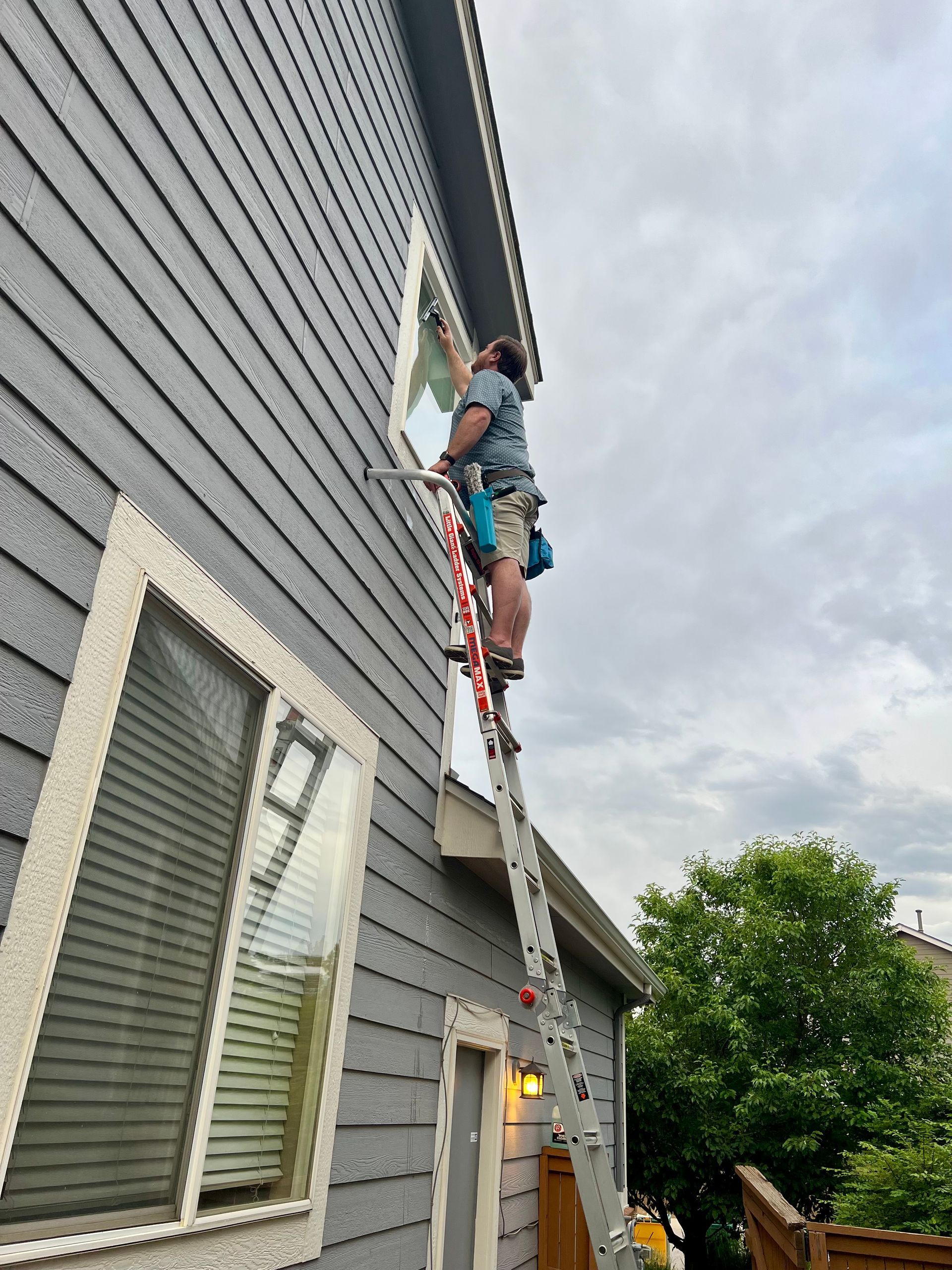 A man is standing on a ladder cleaning a window on the side of a house.