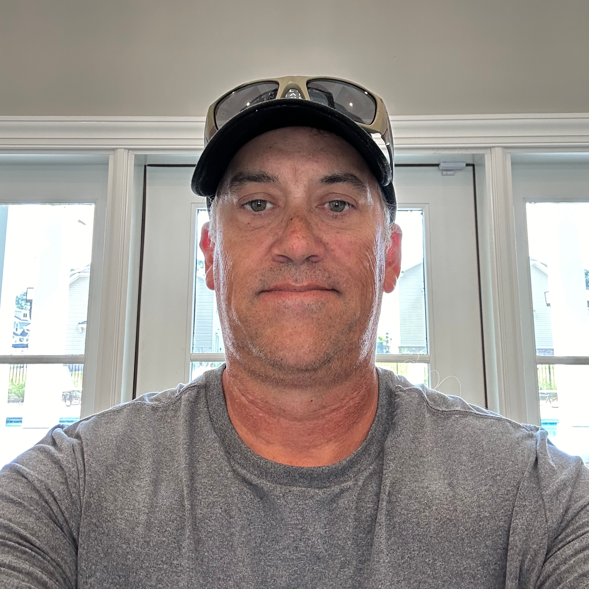 A man wearing a hat and sunglasses is taking a selfie in front of a window.