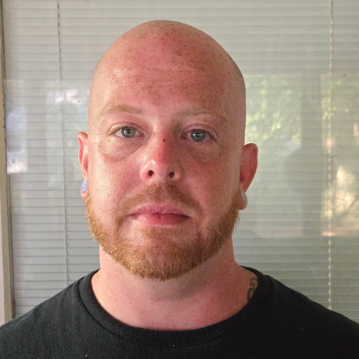 A bald man with a beard is wearing a black shirt and looking at the camera.