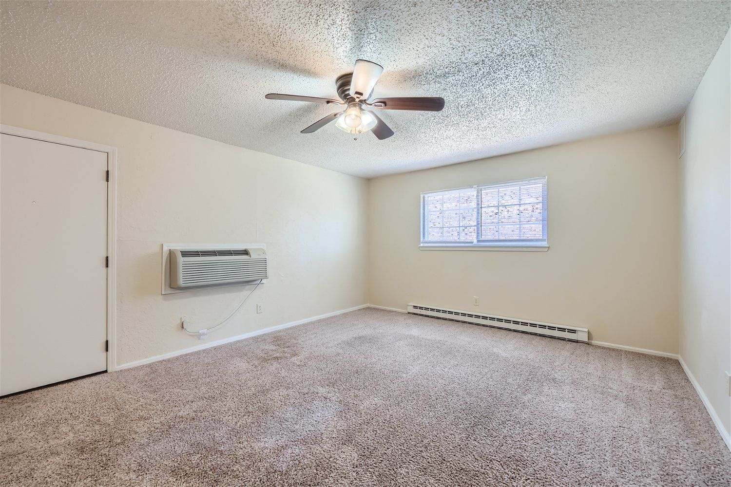 This Copperstone Point apartment room has beige walls, carpet, ceiling fan, window, wall AC unit, and baseboard heater.