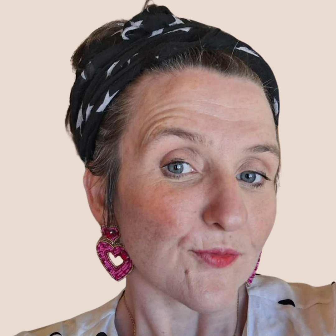 Woman with blue eyes and a black bandana smiles, wearing heart-shaped pink earrings, with a light background.