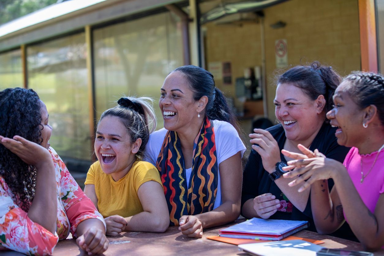 Group of PBS Practitioners laughing together at an outdoor table. Sunlight, colorful clothing, and notebooks are visible.