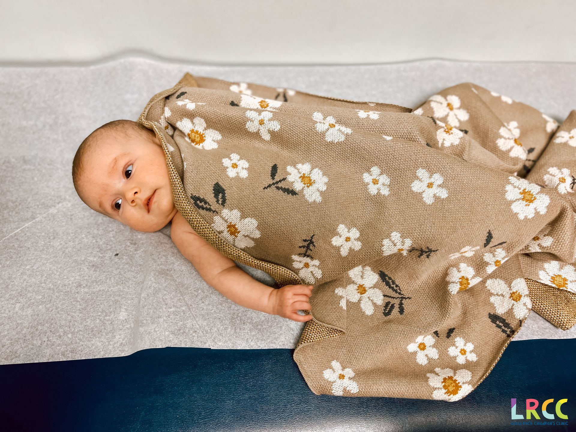 Baby wrapped in a tan floral blanket, looking alert on a medical exam table.