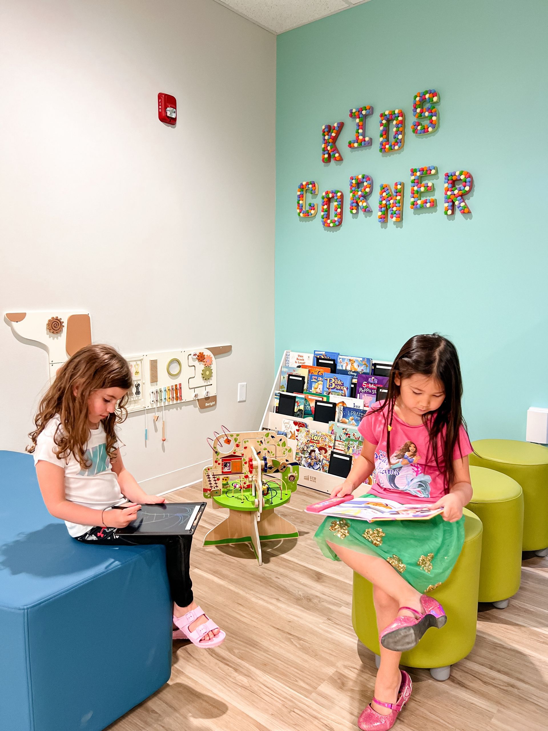 Two girls read in a kids' corner. One uses a tablet, the other a book. Pastel walls and colorful accents.