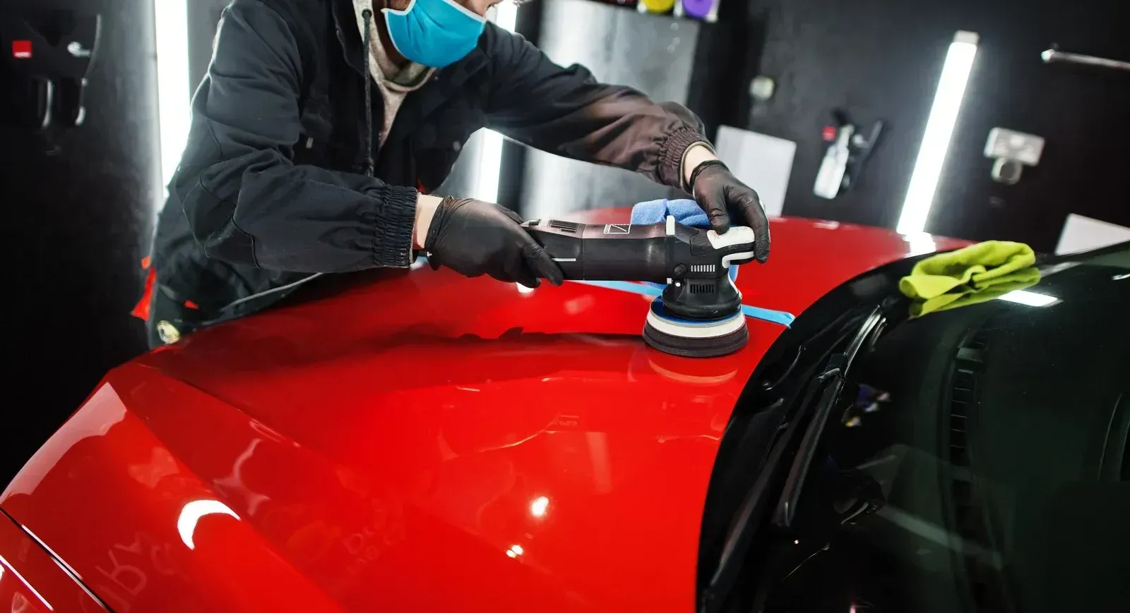 Person polishing a bright red car hood with a machine polisher in a well-lit shop.