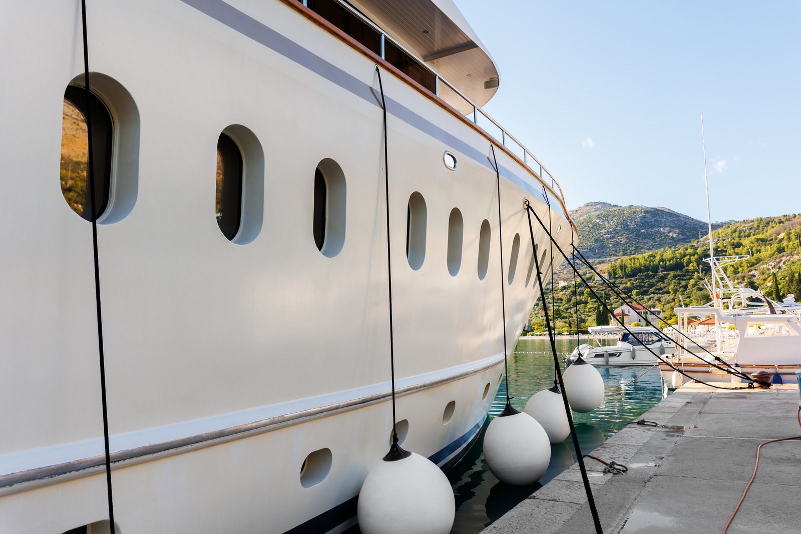 White yacht docked at a marina, secured by black ropes and white buoys; mountains in background.