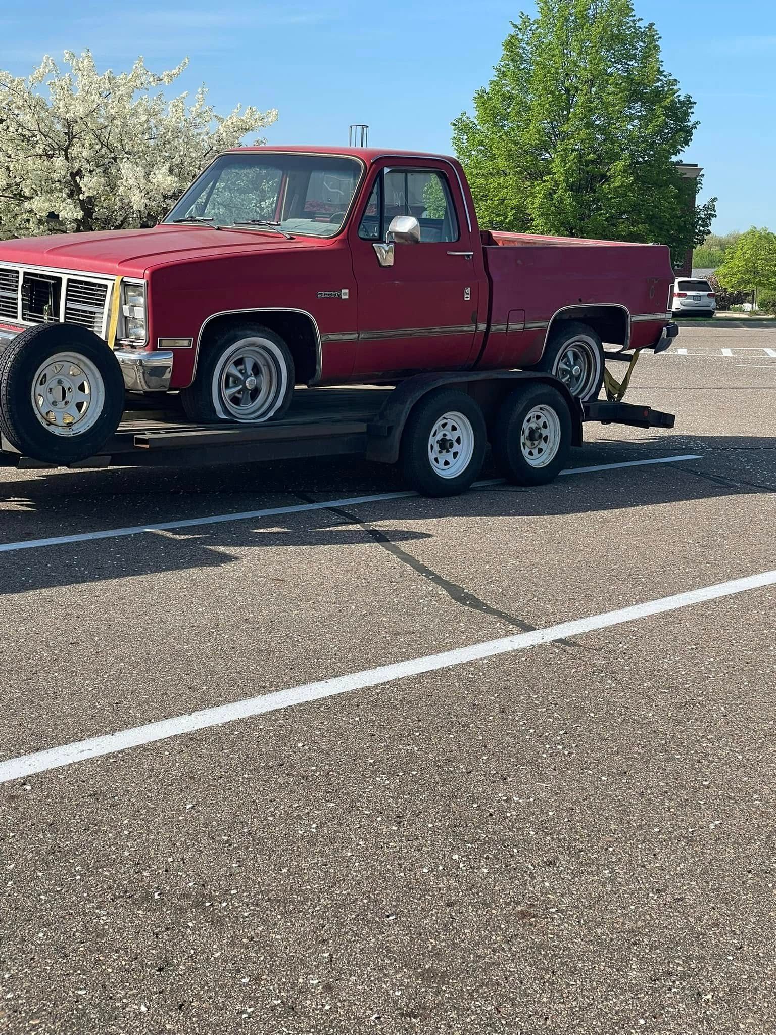 A red truck is being towed by a trailer in a parking lot.
